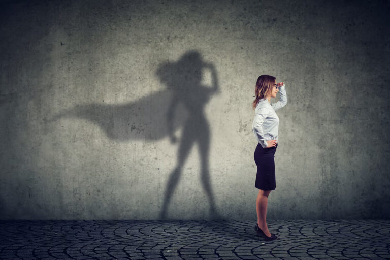 A business woman stands sideways, her shadow casts a figure wearing a superhero cape.